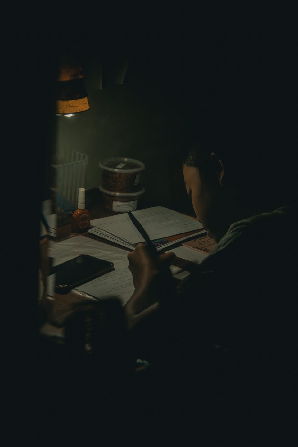 Person writing in notebook at a desk with a lamp.