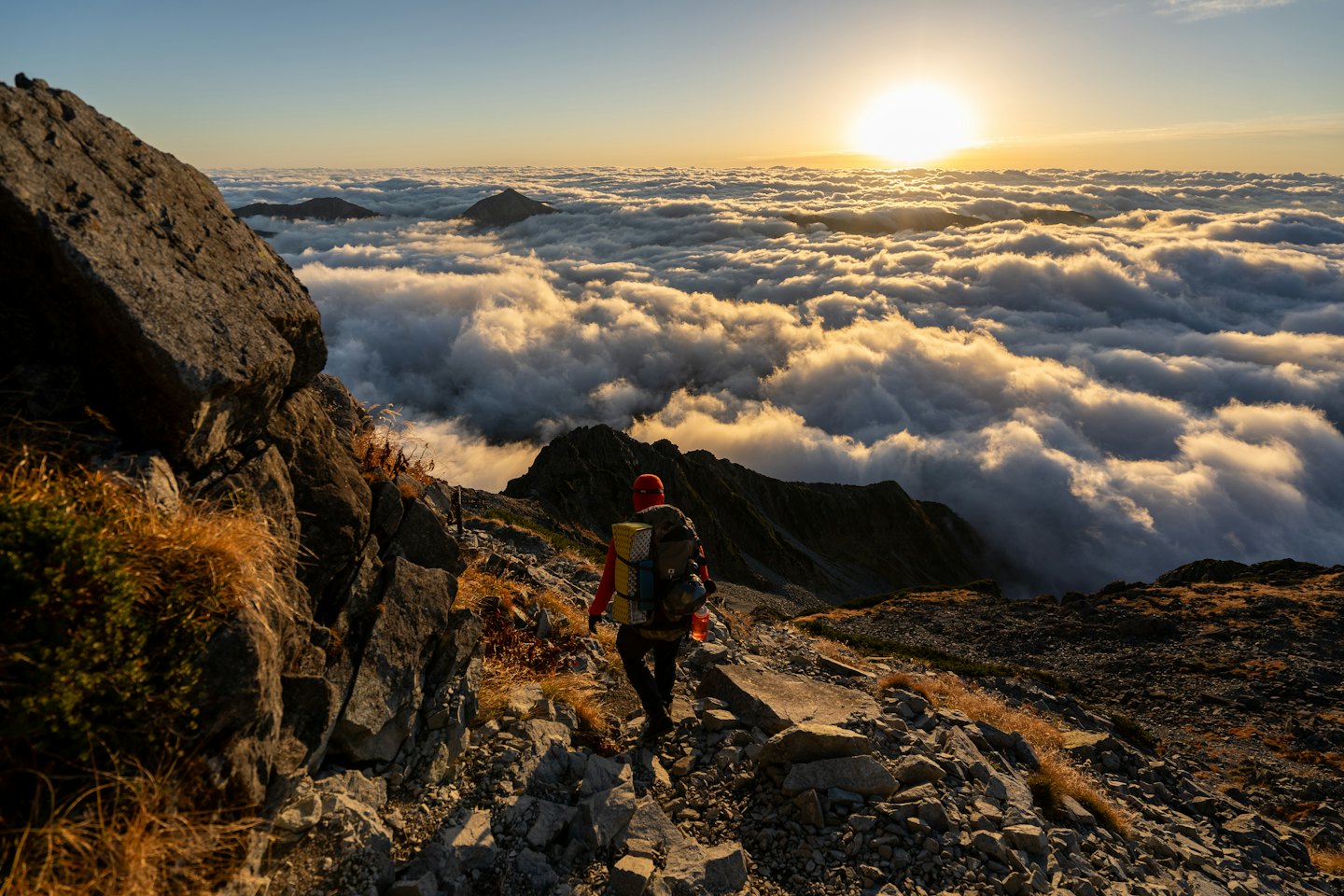 Hiker with red cap walks on rocky mountain trail at sunrise.
